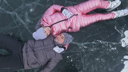 Couple has fun winter walk against background of ice of frozen lake. Lovers lie on clear ice with cracks have fun kiss and hug. View from above. Happy people on snow covered ice. Honeymoon love story.