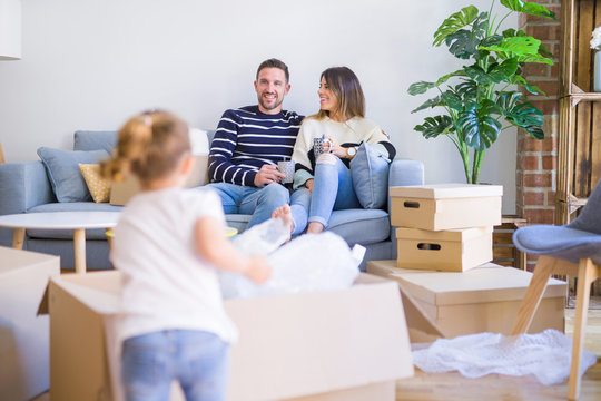 Beautiful family, parents sitting on the sofa drinking coffee looking his kid playing at new home around cardboard boxes