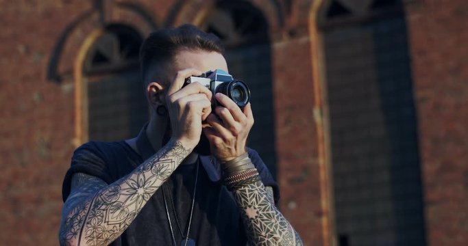 Close up of the Caucasian young hipster male photographer with tattoos taking photo with photocamera at the brick wall background. Outside.