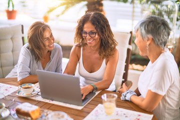 Meeting of middle age women having lunch and drinking coffee. Mature friends smiling happy using laptop at home on a sunny day