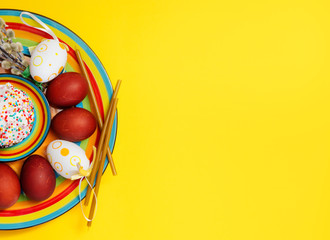 happy Easter. Greeting Easter background with a plate, painted Easter eggs and willow twigs. Concept of Easter banner greeting card on a bright yellow background top view