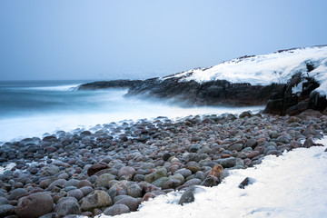 Long exposure of surf breaking on snow covered rocky shores and large stone boulders in the Polar region above the Arctic Circle