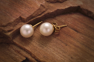 A close up of a pair of white pearl earring studs with yellow gold post and nut on a sandstone background.