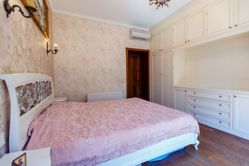 A bedroom in the Provence style. A large white bed with curved legs and back and a pink bedspread. Yellowish Wallpaper with a pattern.