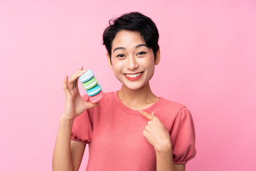 Young Asian girl over isolated pink background holding colorful French macarons with surprise expression