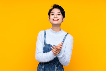 Young Asian girl in overalls over isolated yellow background applauding