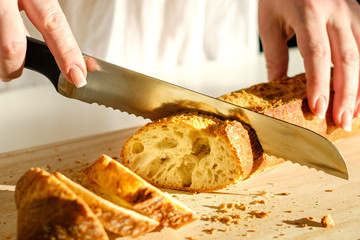 Slicing tasty, fresh, crisp bread. Beautiful female hands, a knife for cutting bread and sliced baguette closeup. Floury product on a cutting board. Healthy eating concept.