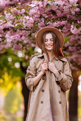 Portrait of young beautiful fashionable girl in hat posing near blooming tree with pink flowers on a sunny day. Spring, girl near a flowering sakura tree. Female beauty and fashion. City lifestyle.