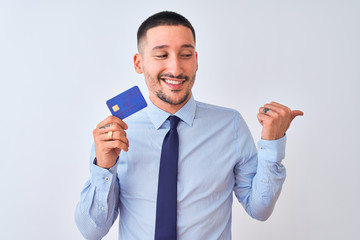 Young business man holding credit card over isolated background pointing and showing with thumb up to the side with happy face smiling