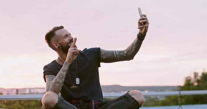 Handsome Caucasian young guy in hipster style sitting on the roof top over the town and taking selfie photo with phone camera.