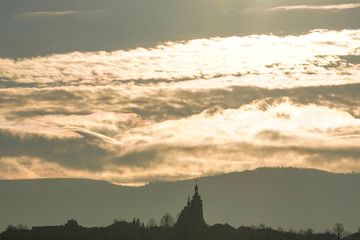 Obraz premium Majestic yellow sunset in the mountains landscape. Dramatic sky clouds, partly clear.sun hides in clouds,illuminating. mountains with forests on hills,a Church domes and roofs on horizon. evening time