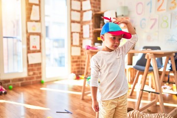 Beautiful blonde toddler wearing fanny colorful propeller cap. Standing around lots of toys at kindergarten