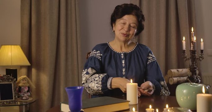 Portrait of old Caucasian woman in blue blouse sitting at the table with candles and shuffling cards. Smiling witch putting cards on table and talking. Foretelling, augury, mystic. Cinema 4k ProRes HQ