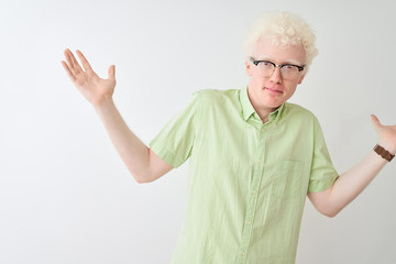 Young albino blond man wearing green shirt and glasses over isolated white background clueless and...