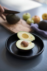Avocado cut in half on dark plate, , blue table with other avocado being cut on background.