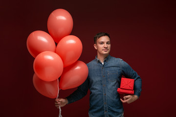 Handsome man with coral balloons and red giftboxes isolated over dark red background.