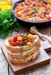 Spanish vegetable dish pisto manchego made of tomatoes, zucchini, bell peppers, onions and eggplant served on white bread. Wooden background. Selective focus