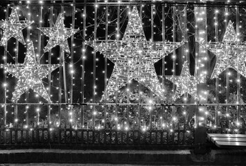 Abstract background. Black and  white Christmas background with shiny stars and glowing Christmas lights. Ornate fence with light garlands. Blurred backdrop for winter holiday. New Year decorations