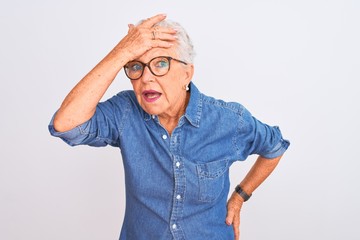 Senior grey-haired woman wearing denim shirt and glasses over isolated white background surprised with hand on head for mistake, remember error. Forgot, bad memory concept.