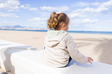 Fototapeta na wymiar Beautiful toddler child girl wearing jacket sitting on the bench in the promenade