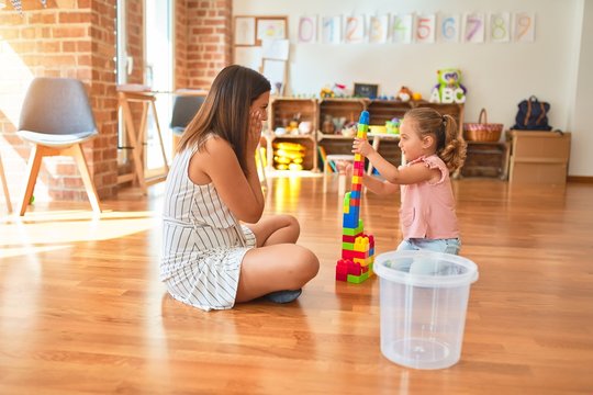 Beautiful teacher and blond toddler girl building tower using plastic blocks at kindergarten