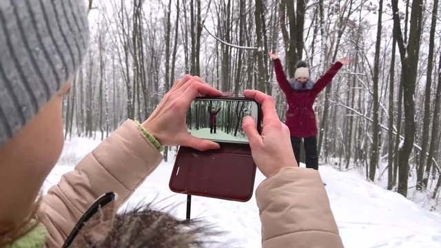 Mother taking photos of her teenager daughter girl on smartphone and female posing for a photo in the Russian winter snowy forest. Healthy open air spending holidays, weekend outdoors in cold weather