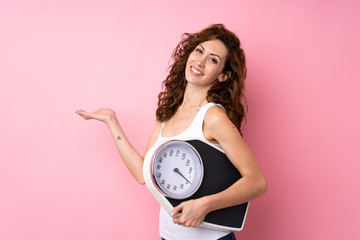 Young woman with curly hair holding a weighing machine over isolated pink background