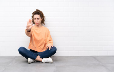 Fototapeta premium Young woman with curly hair sitting on the floor making stop gesture with her hand