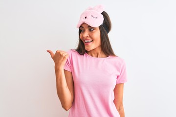Obraz na płótnie Canvas Young beautiful woman wearing sleep mask standing over isolated white background smiling with happy face looking and pointing to the side with thumb up.