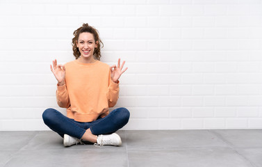 Young woman with curly hair sitting on the floor showing an ok sign with fingers