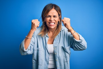 Middle age beautiful woman wearing casual shirt standing over isolated blue background angry and mad raising fists frustrated and furious while shouting with anger. Rage and aggressive concept.