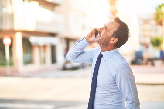 Middle age handsome businessman standing on the street talking on the smartphone smiling