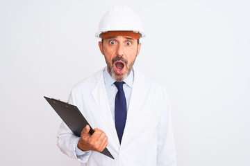 Senior engineer man wearing security helmet holding clipboard over isolated white background scared in shock with a surprise face, afraid and excited with fear expression