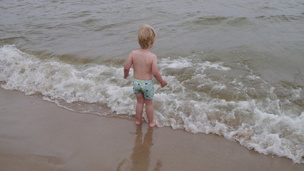 Chłopiec bawi się na plaży nad morzem w piasku robiąc wielki zamek z piasku i wchodzi do wody. Wakacyjny wyjazd z dzieckiem w lato podczas urlopu. - obrazy, fototapety, plakaty