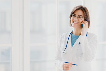 Shot of pleased female physician or surgeon gives prescription via mobile phone, has conversation with patient, holds pen, wears white uniform, works in hospital, stands indoor against window