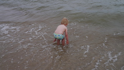 Chłopiec bawi się na plaży nad morzem w piasku robiąc wielki zamek z piasku i wchodzi do wody. Wakacyjny wyjazd z dzieckiem w lato podczas urlopu. - obrazy, fototapety, plakaty