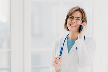 Positive woman doctor consults patient via cellphone, looks gladfully at camera, holds pen, wears...