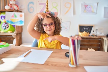 Beautiful toddler wearing glasses and unicorn diadem sitting on desk at kindergarten smiling making frame with hands and fingers with happy face. Creativity and photography concept.