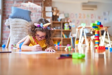 Beautiful toddler wearing glasses and unicorn diadem lying down on the floor drawing using paper and pencil at kindergarten