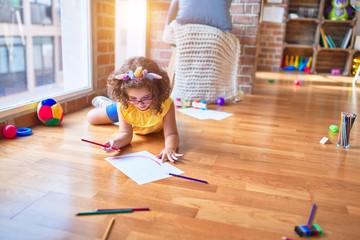 Beautiful toddler wearing glasses and unicorn diadem lying down on the floor drawing using paper and pencil at kindergarten © Krakenimages.com