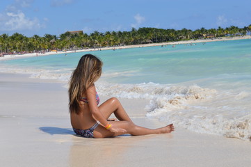 Fototapeta na wymiar Pretty young woman with highlights is sitting on white sand beach and looking at a waves in Riviera Maya, Mexico. 