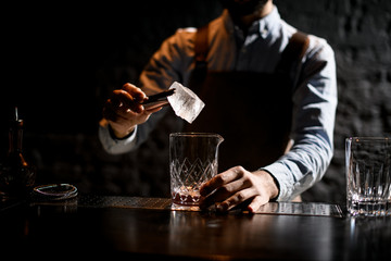 Professional male bartender putting ice cube with twezzers to the glass
