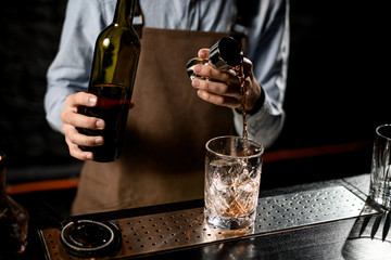 Professional bartender pouring a brown alcoholic drink from the steel jigger to a glass