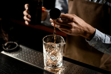 Professional male bartender pouring a brown alcoholic drink from the steel jigger to a glass cup