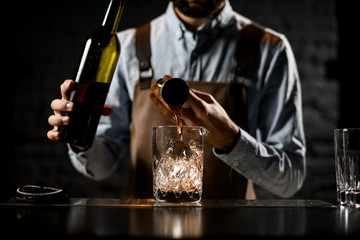 Professional bartender pouring a golden alcoholic drink from the steel jigger to a glass cup