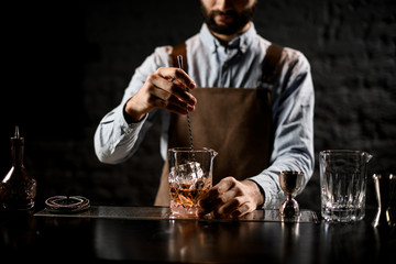 Male bartender stirring a brown cocktail with ice cubes in the glass with special spoon