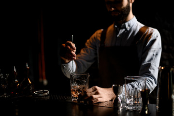 Male bartender stirring a golden cocktail with ice cubes in the glass with special spoon