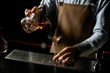 Male bartender pouring a brown alcoholic drink from the measuring cup with straner to a martini glass