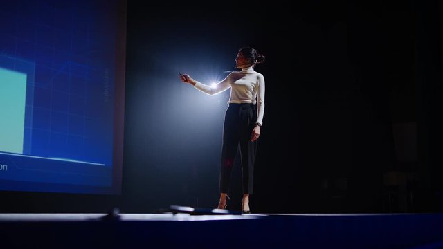 On Stage, Successful Female Speaker Presents Technological Product, Uses Remote Control for Presentation, Showing Infographics, Statistics Animation on Screen. Live Event / Device Release. Slow Motion