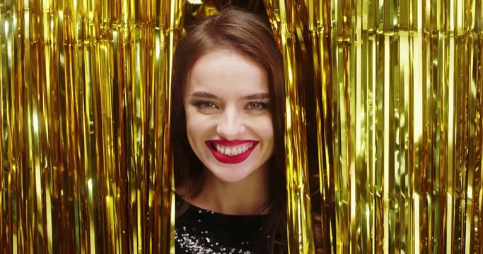 Close up of the charming and pretty young brunette woman looking out from the golden sparkle confetty strips and smiling cheerfully on the New Year Eve. Portrait.